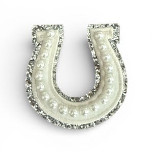 May include: A white horseshoe-shaped embellishment with a row of faux pearls and a silver glitter outline. The horseshoe is a symbol of good luck, often used in crafts and jewellery. The item is suitable for various decorative purposes.