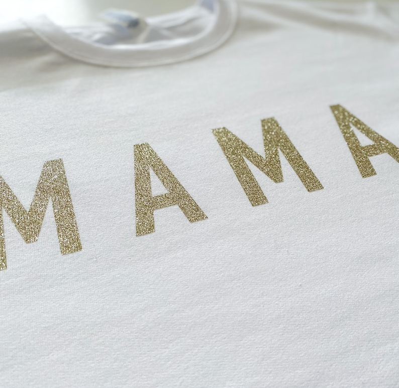 May include: White t-shirt with the word "MAMA" in gold glitter lettering.