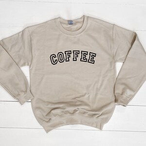 K&ouml;nnte beinhalten: Beiges Sweatshirt mit dem Wort "COFFEE" in schwarzen, umrandeten Buchstaben auf der Brust. Das Sweatshirt hat einen Rundhalsausschnitt und lange &Auml;rmel. Der Hintergrund ist eine wei&szlig;e Holzoberfl&auml;che.