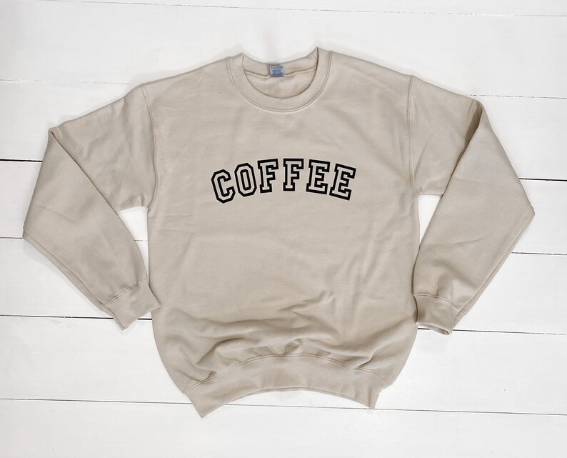 K&ouml;nnte beinhalten: Beiges Sweatshirt mit dem Wort "COFFEE" in schwarzen, umrandeten Buchstaben auf der Brust. Das Sweatshirt hat einen Rundhalsausschnitt und lange &Auml;rmel. Der Hintergrund ist eine wei&szlig;e Holzoberfl&auml;che.