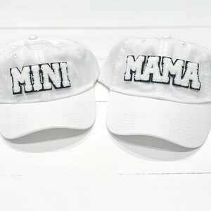 May include: Two white baseball caps with black trim. One cap says "MINI" and the other says "MAMA".