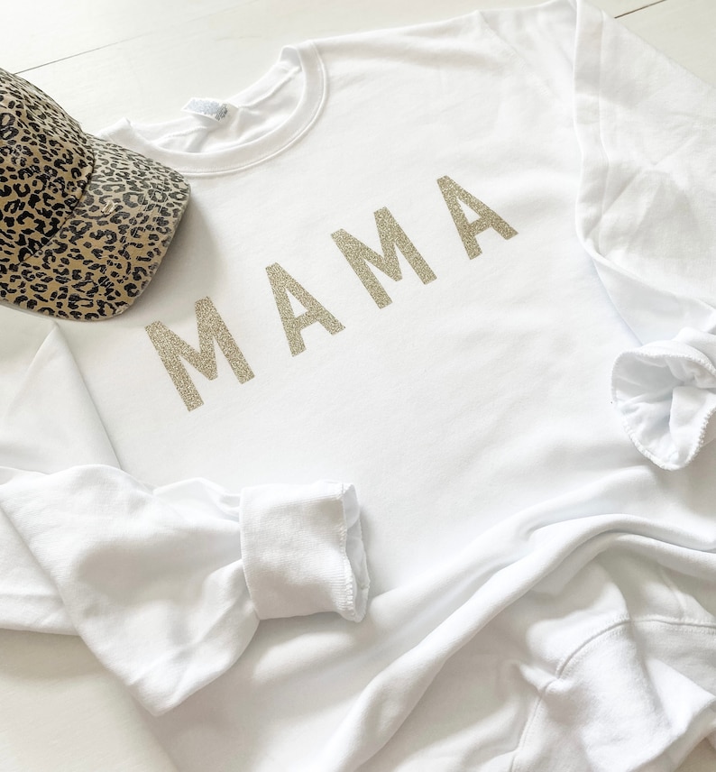May include: White sweatshirt with long sleeves and a crew neck. The word "MAMA" is printed in gold glitter on the front of the sweatshirt.
