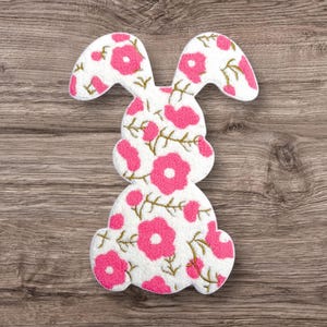 May include: A white bunny-shaped applique with a floral pattern in pink and gold. The bunny has large ears and is sitting upright. The applique is made of a soft, textured material and is likely for crafts or decoration.