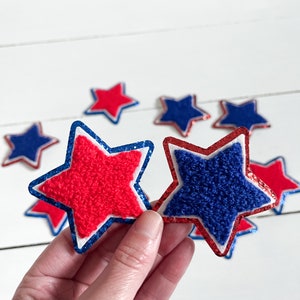 May include: Red and blue felt star-shaped patches with white borders and glitter accents. The stars are held in a hand.
