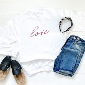 May include: White sweatshirt with the word "love" written in red cursive script. The sweatshirt is laid flat on a white wooden surface with a pair of black fuzzy slippers, a pair of blue jeans, and a leopard print headband.