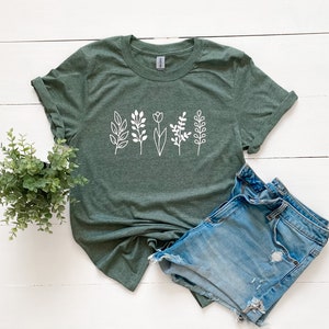 May include: A green t-shirt featuring a white graphic of stylised flowers and leaves. The shirt is laid flat on a white wooden surface with a pair of blue denim shorts.