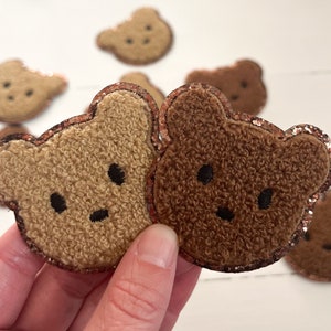 May include: Two fuzzy brown teddy bear patches with glitter trim. The patches are held in a hand.