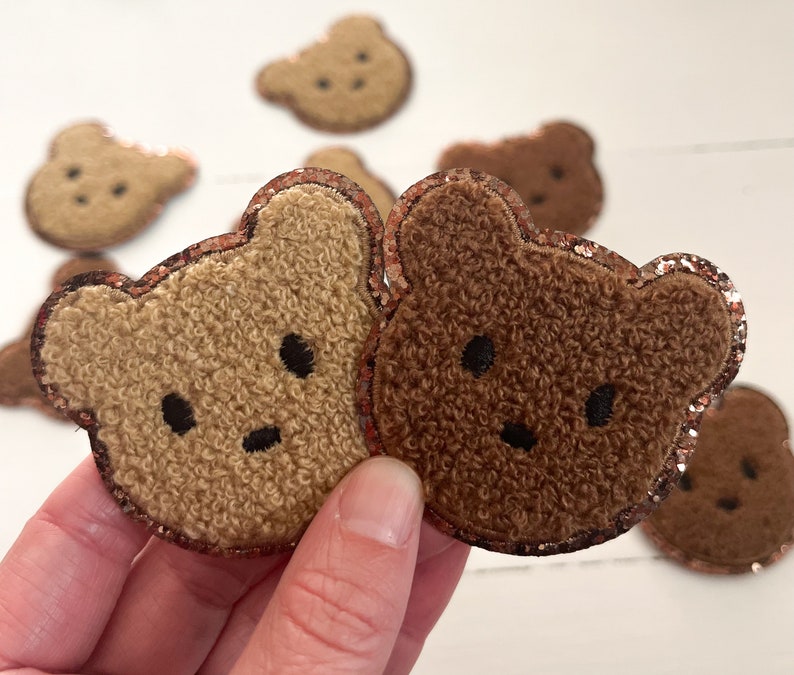 May include: Two fuzzy brown teddy bear patches with glitter trim. The patches are held in a hand.