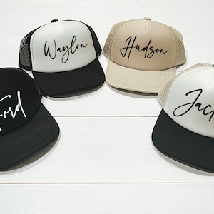 May include: Four trucker hats with black mesh backs and different coloured fronts. The hats are personalised with the names "Waylon", "Hudson", "Ford", and "Jack" in a cursive font.