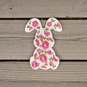 May include: Embroidered bunny patch with a white background and pink floral design. The bunny shape is outlined in white, and the flowers have gold-colored stems and leaves. The patch is likely for decorative purposes.