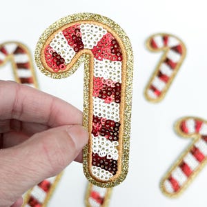 May include: A candy cane patch with red, white, and burgundy sequins, outlined with gold glitter. The patch is held by a hand, with other candy cane patches in the background. A festive Christmas decoration.