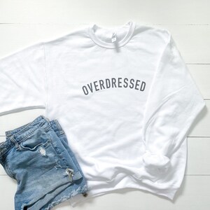 May include: White sweatshirt with the text "OVERDRESSED" printed in gray on the front. The sweatshirt is paired with blue denim shorts.