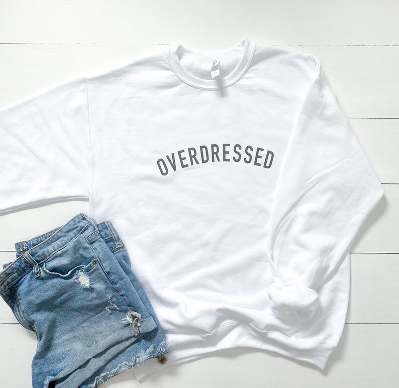 May include: White sweatshirt with the text "OVERDRESSED" printed in gray on the front. The sweatshirt is paired with blue denim shorts.