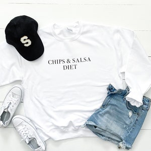 May include: White sweatshirt with the text "Chips & Salsa Diet" printed on the front. The sweatshirt is laid out on a white wooden surface with a black baseball cap, blue denim shorts, and white trainers.