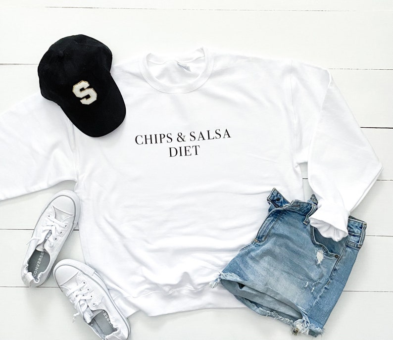 May include: White sweatshirt with the text "Chips & Salsa Diet" printed on the front. The sweatshirt is laid out on a white wooden surface with a black baseball cap, blue denim shorts, and white trainers.