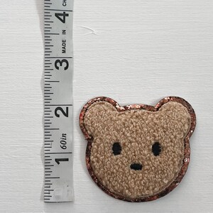 May include: A brown fuzzy bear applique with a copper glitter outline. The bear has black eyes and a black nose.