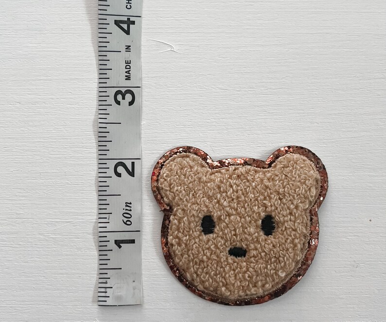 May include: A brown fuzzy bear applique with a copper glitter outline. The bear has black eyes and a black nose.