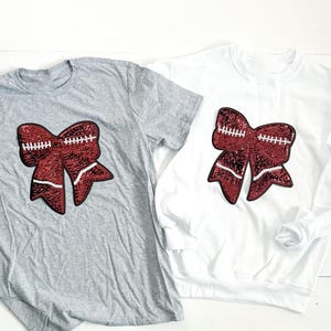 May include: A gray t-shirt and a white sweatshirt, each featuring a large, sequined bow design. The bow is a deep red color with white stitching, resembling a football. The garments are displayed on a white wooden surface.