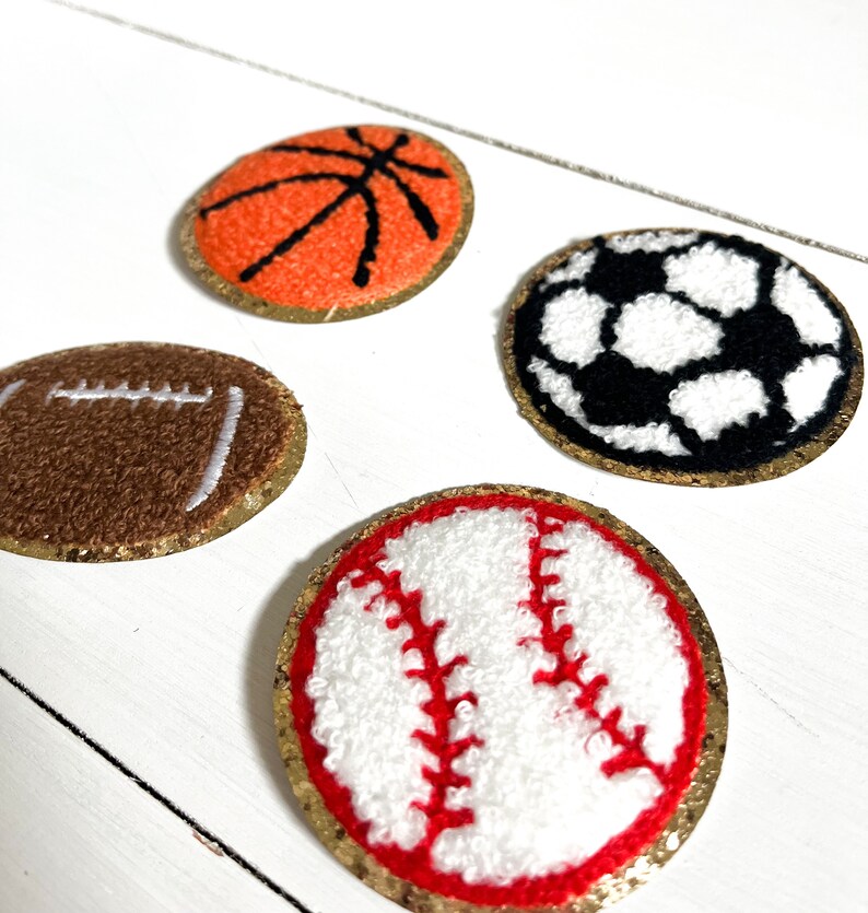 May include: Four felt sports ball patches with gold glitter trim. The patches are of a basketball, soccer ball, football, and baseball.