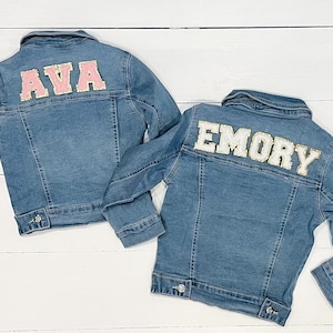 May include: Two denim jackets with personalized names on the back. The jacket on the left has the name "AVA" in pink letters with gold trim. The jacket on the right has the name "EMORY" in white letters with gold trim.
