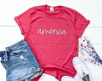 Puede incluir: Una camiseta roja con la palabra "america" escrita en letras blancas cursivas. La camiseta se combina con pantalones cortos de mezclilla y zapatillas blancas.