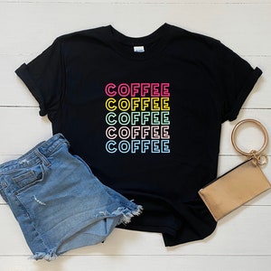 May include: Black t-shirt with the word "COFFEE" repeated five times in a rainbow color pattern. The shirt is laid flat on a white wooden surface with a pair of blue denim shorts and a gold clutch.