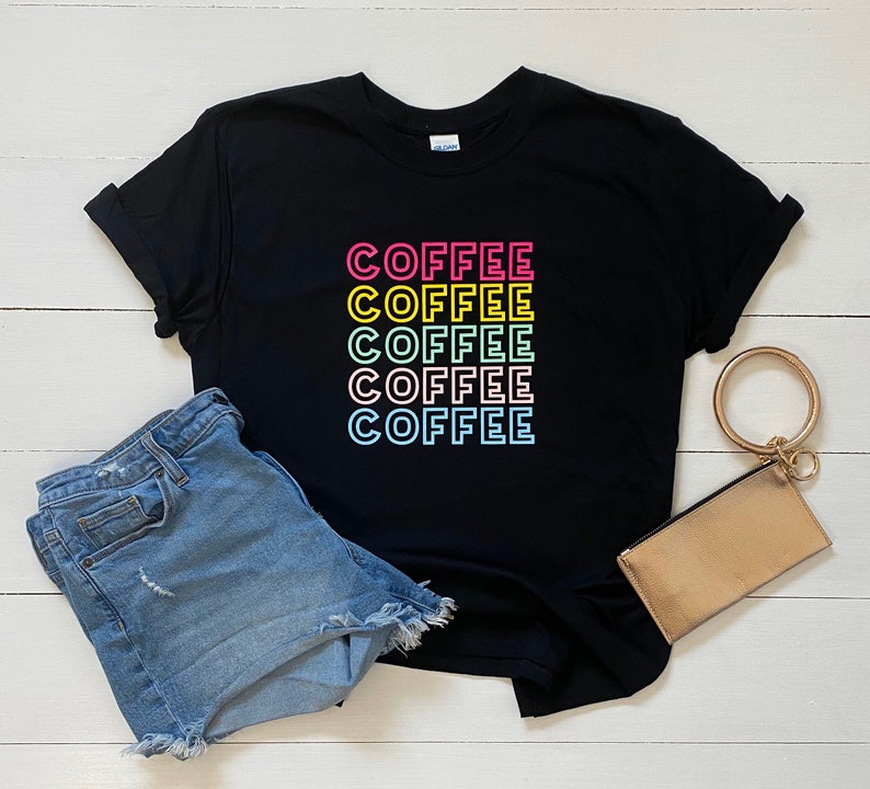 May include: Black t-shirt with the word "COFFEE" repeated five times in a rainbow color pattern. The shirt is laid flat on a white wooden surface with a pair of blue denim shorts and a gold clutch.