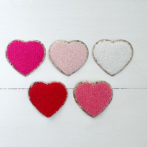 May include: Five felt heart-shaped patches with gold trim. The hearts are pink, white, red, and two shades of pink.
