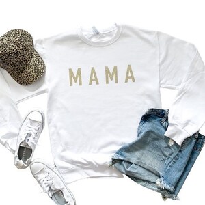 May include: White long-sleeved sweatshirt with the word "MAMA" in gold glitter lettering. The sweatshirt is paired with denim shorts and a leopard print baseball cap.