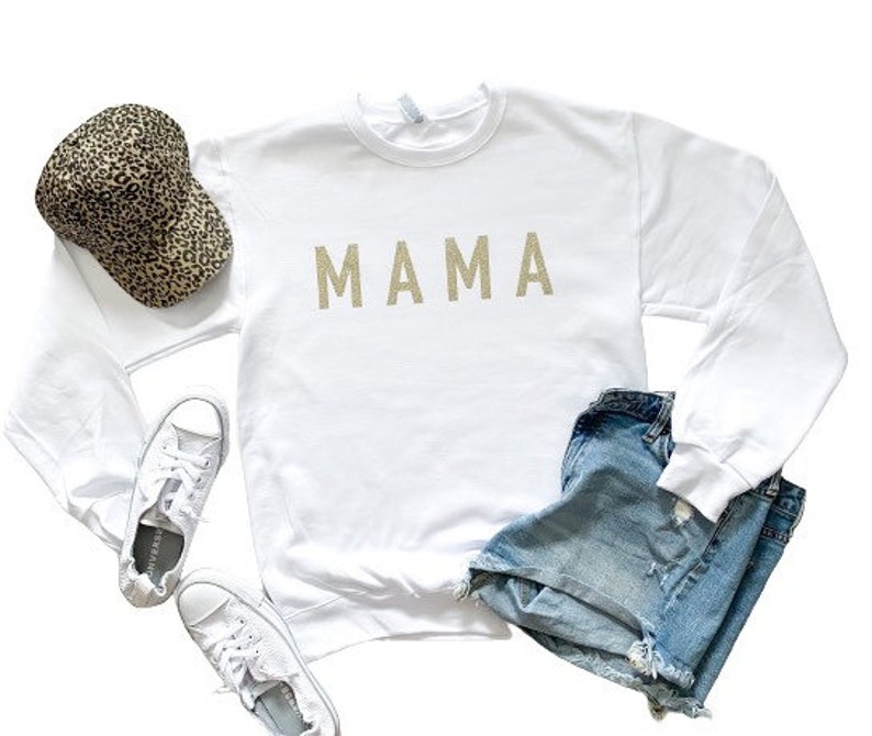 May include: White long-sleeved sweatshirt with the word "MAMA" in gold glitter lettering. The sweatshirt is paired with denim shorts and a leopard print baseball cap.
