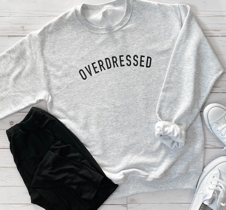 May include: A gray sweatshirt with the text "OVERDRESSED" printed in black on the front. The sweatshirt is folded and laid on a white wooden surface.