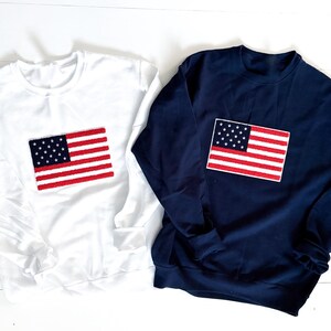 May include: Two sweatshirts, one white and one navy blue, each featuring an embroidered American flag on the front.