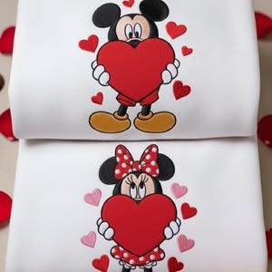 Può includere: Due felpe bianche, una sopra l'altra. La felpa superiore presenta Topolino che tiene un cuore rosso, circondato da cuori rossi. La felpa inferiore mostra Minnie che tiene un cuore rosso, circondata da cuori rosa. Tema romantico.