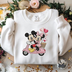 May include: White sweatshirt featuring a colorful graphic of Mickey and Minnie Mouse riding a pink scooter. Minnie wears a pink polka-dot dress and bow, while Mickey wears red shorts. Heart-shaped balloons and a basket complete the design.