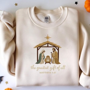 May include: Cream-colored sweatshirt featuring an embroidered Nativity scene with Mary, Joseph, and baby Jesus. The design includes a star and the text "the greatest gift of all" with the Bible verse Matthew 1:21.