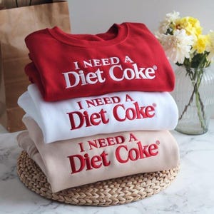 Könnte beinhalten: Drei übereinander gestapelte Sweatshirts in Rot, Weiß und Beige. Jedes Sweatshirt hat den gestickten Text "I NEED A Diet Coke" in einer Kontrastfarbe. Die Sweatshirts liegen auf einem geflochtenen Untersetzer.