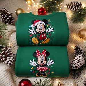 Op de afbeelding: Twee groene sweatshirts, de ene bovenop de andere. Het bovenste sweatshirt heeft Mickey Mouse met een kerstmuts en kerstverlichting. Het onderste sweatshirt heeft Minnie Mouse met een rode polka-dot strik en jurk, ook met lichtjes.
