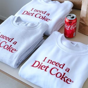 Puede incluir: Sudaderas blancas y grises con la frase bordada "I need a Diet Coke" en rojo. También hay una lata roja de Diet Coke. Las sudaderas están cuidadosamente apiladas, lo que sugiere una prenda de vestir.