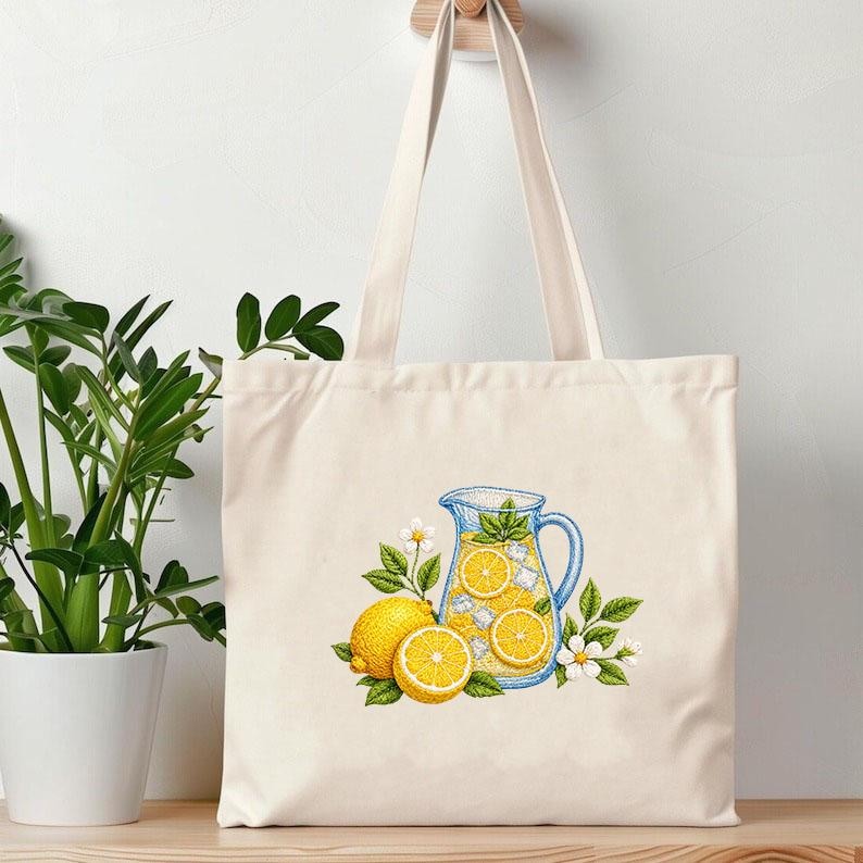 May include: Cream-colored tote bag featuring an embroidered design of a pitcher of lemonade with ice cubes, lemon slices, and mint leaves. Two whole lemons and a lemon half are also depicted, along with white flowers and green leaves. The bag has long, cream-colored handles.
