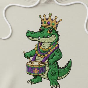 May include: Embroidered design of a green alligator wearing a gold crown and Mardi Gras beads, playing a drum with a purple fleur-de-lis. The design is on a light beige hoodie.