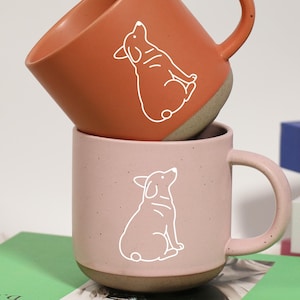 Puede incluir: Dos tazas de cerámica, una terracota y otra rosa, apiladas una encima de la otra. Cada taza presenta un contorno blanco de un perro. Las tazas están sobre un libro verde, con otros libros en el fondo.