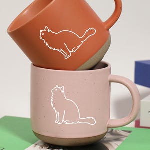 Może przedstawiać: Dwa ceramiczne kubki, jeden ułożony na drugim. Górny kubek jest pomarańczowy z białą sylwetką kota, a dolny kubek jest różowy z białą sylwetką kota. Kubki stoją na zielonej książce.