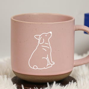May include: A pink ceramic mug with a white outline of a dog. The mug has a brown base and a curved handle. The mug is sitting on a wooden coaster and a fluffy white surface.