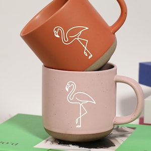 Puede incluir: Dos tazas de cerámica, una terracota y otra rosa, apiladas una encima de la otra. Cada taza presenta un contorno de flamenco blanco. Las tazas están sobre un libro verde, con otros libros en el fondo.