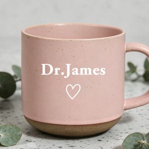 Può includere: Una tazza in ceramica rosa con la scritta "Dr. James" e un simbolo a cuore. La tazza ha una base marrone e un manico. La tazza è su una superficie maculata con foglie verdi.