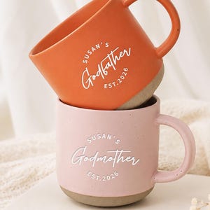 Puede incluir: Una pila de dos tazas de cerámica con acabado mate. La taza superior es naranja con el texto blanco Susan's Godfather EST. 2026. La taza inferior es de color rosa claro con motas y el texto blanco Susan's Godmother EST. 2026. Ambas tienen asas.