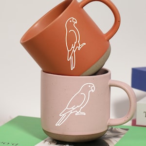 May include: Two ceramic mugs, one terracotta and one pink, stacked on top of each other. Each mug features a white outline of a parrot. The mugs are resting on a green book, with other books visible in the background.