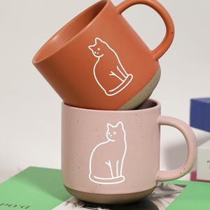 Puede incluir: Dos tazas de cerámica, una apilada encima de la otra. La taza superior es naranja y la inferior es rosa. Cada taza presenta un contorno blanco de un gato. Las tazas están sobre un libro verde.