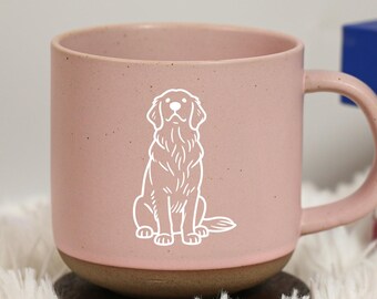May include: A pink ceramic mug with a white outline of a dog. The mug has a brown base and a curved handle. The mug is sitting on a wooden coaster and a fluffy white surface.