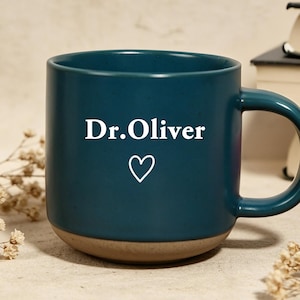 Puede incluir: Una taza de cerámica color verde azulado con el texto "Dr. Oliver" y un símbolo de corazón blanco. La taza tiene una base marrón y un asa grande. Flores secas y libros en el fondo.